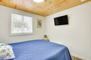 Une chambre avec une couvre-lit bleue, un plafond en bois, un téléviseur fixé au mur et une fenêtre avec des stores.