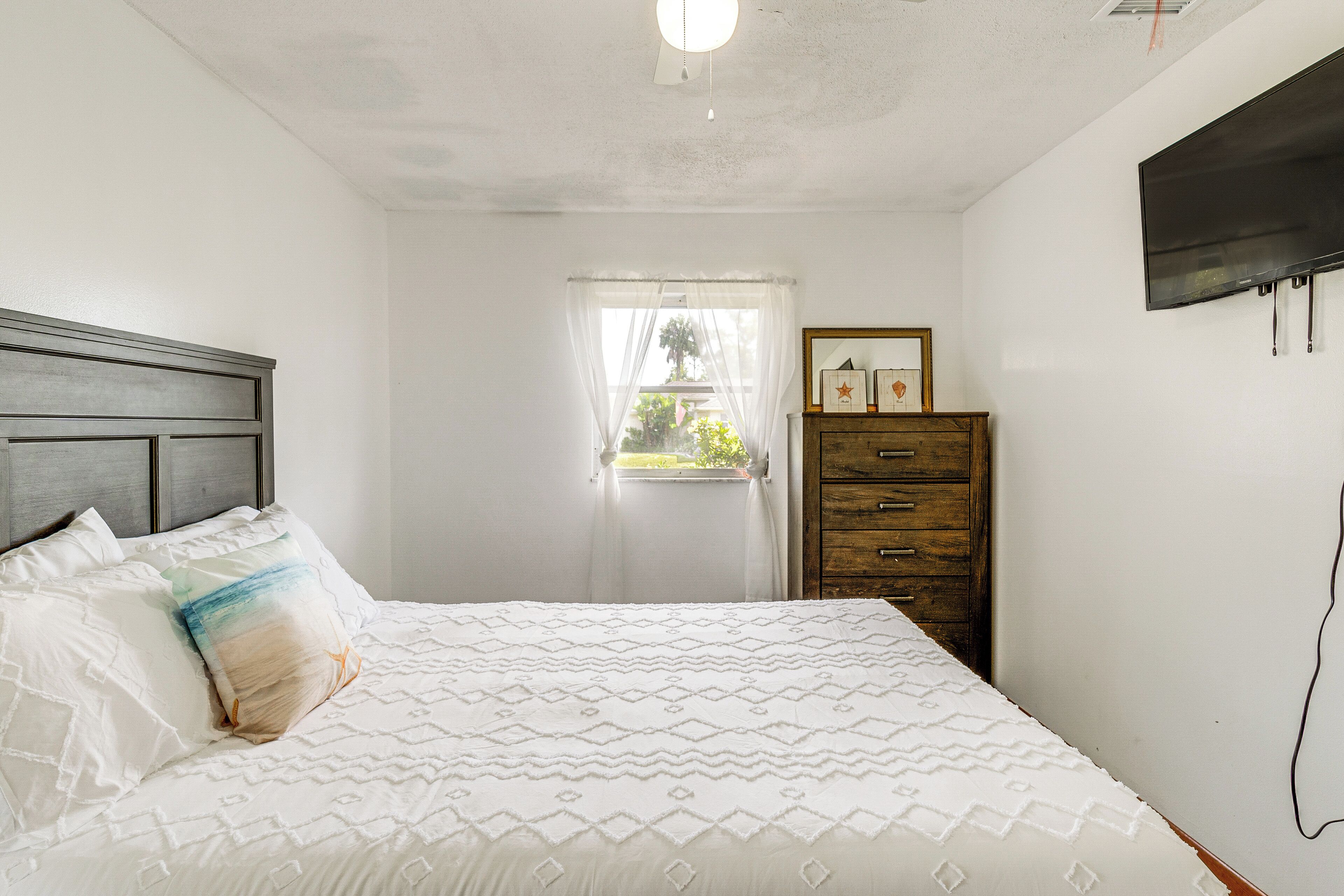 Une chambre à coucher avec un lit, une commode en bois, un téléviseur fixé au mur et une fenêtre ornée de rideaux légers.