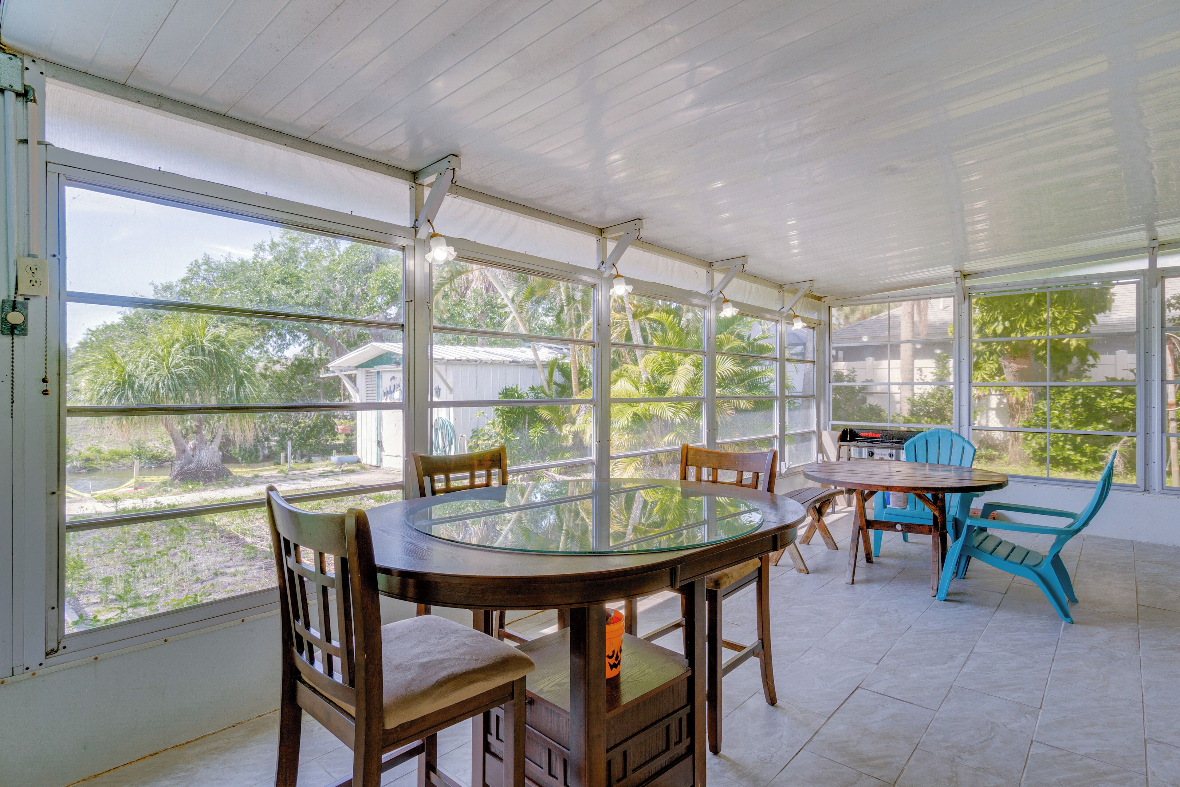 A covered patio with a glass table surrounded by wooden chairs and a blue chair, overlooking a garden with trees and a house in the background.