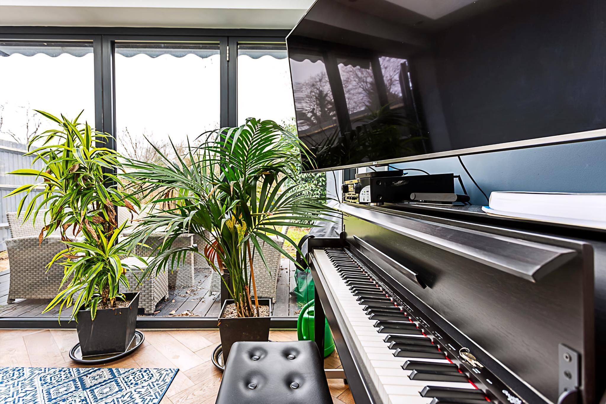 Een piano in een kamer met grote ramen, potplanten en uitzicht op buiten.