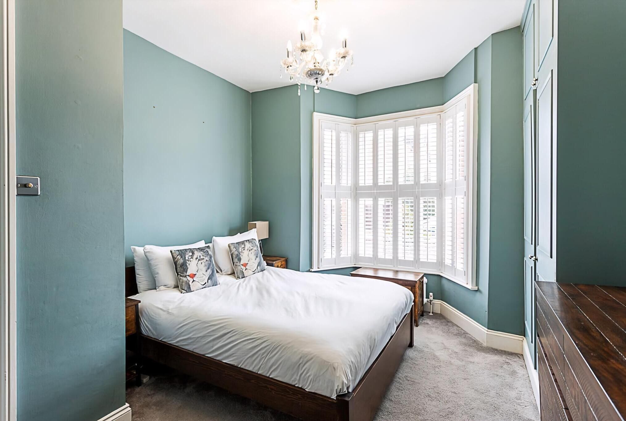 A bedroom with a large bed, a nightstand, a chandelier, and a bay window with shutters.