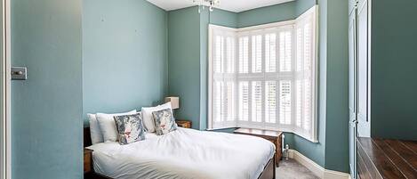 A bedroom with a large bed, a nightstand, a chandelier, and a bay window with shutters.