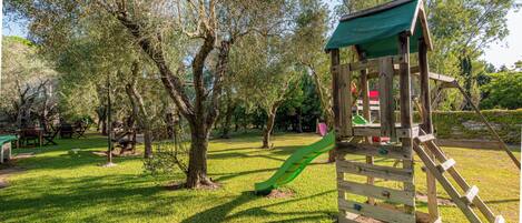 A wooden play structure with a slide and climbing area, surrounded by grass and trees.