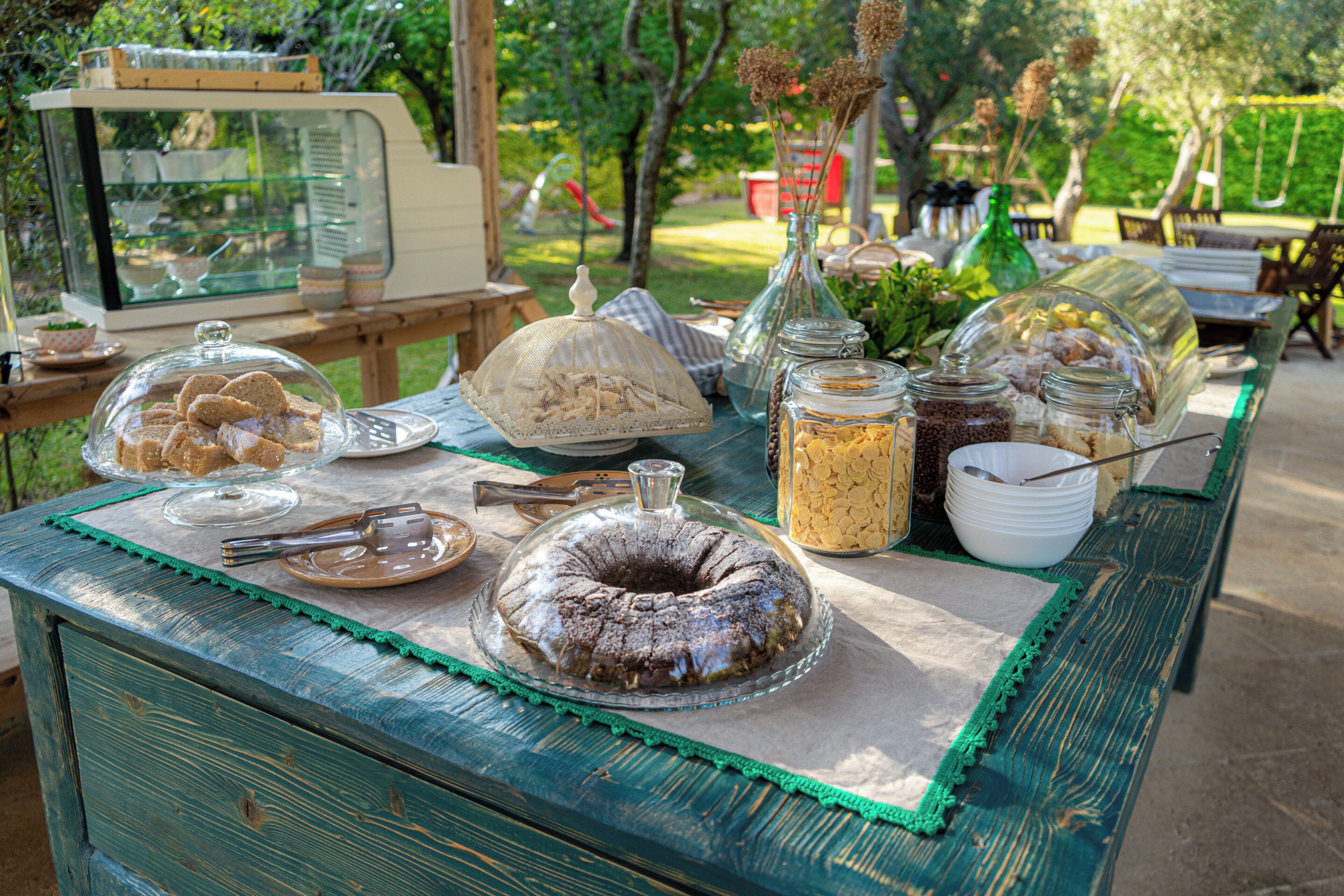Una mesa de bufé con diversos alimentos y un pastel, ubicada al aire libre con una mesa y sillas de madera.