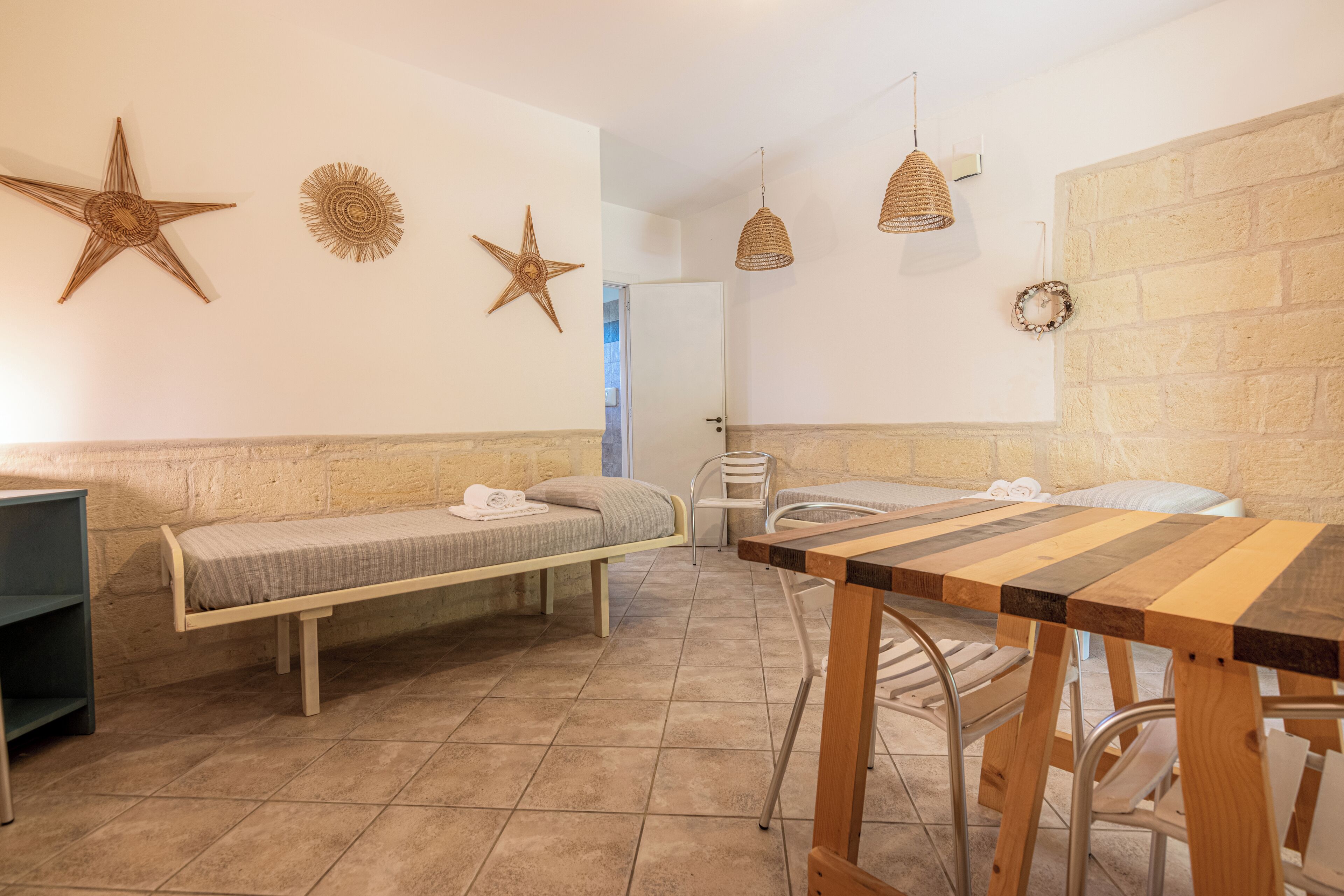 Une petite pièce agréable et chaleureuse, avec deux lits simples, une table en bois et un banc. Les murs sont décorés de motifs en forme d’étoiles, et un luminaire tressé est suspendu au plafond.