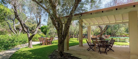 An outdoor patio area with wooden tables and chairs, surrounded by trees and greenery.