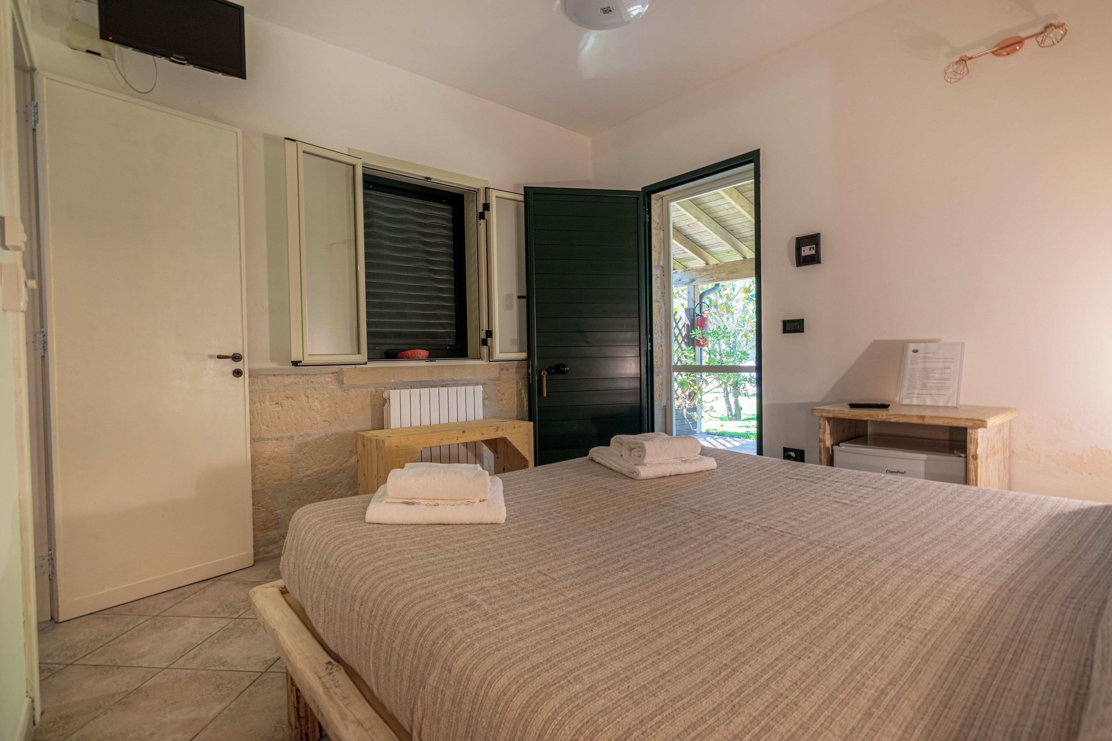 A bedroom with a bed, a wooden bench, a small table with a lamp, a mirror, and a door leading to a balcony.