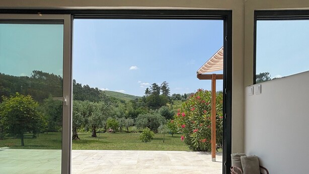 Ein Zimmer mit einer großen Schiebeglasdoor, die auf eine Terrasse mit Blick auf eine grüne Landschaft und einen Hügel führt. Auf dem Boden liegt eine rote Matte, ein blaues Kissen und ein gewebter Korb mit Kissen.