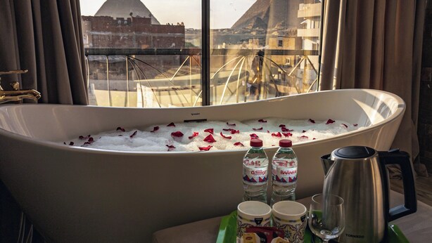 A bathtub filled with ice and rose petals, with bottles of water and a coffee pot on a tray, and a view of pyramids through a window. - Jimmy Pyramids Hotel  (Giza)