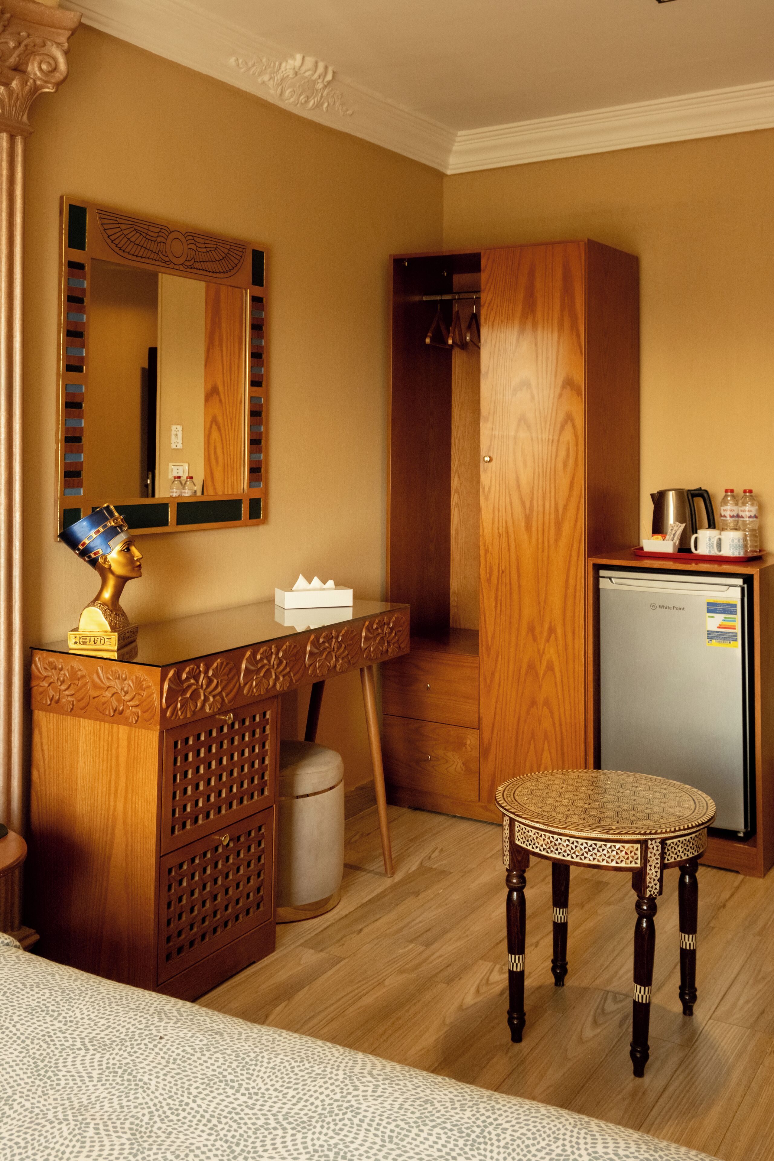 A hotel room with a wooden vanity, a small refrigerator, a round wicker table, and a decorative bust on the vanity.