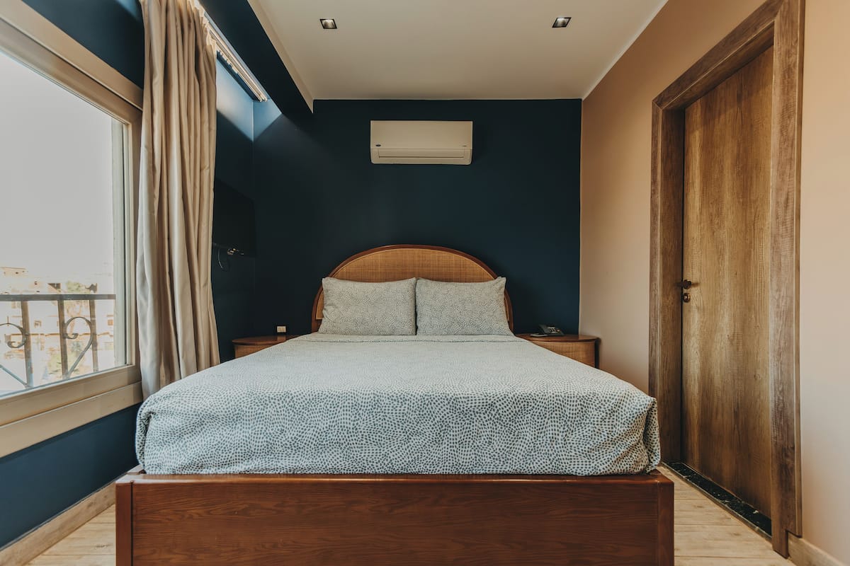 A bedroom with a bed, bedside tables, a window with curtains, an air conditioning unit, and a wooden door.