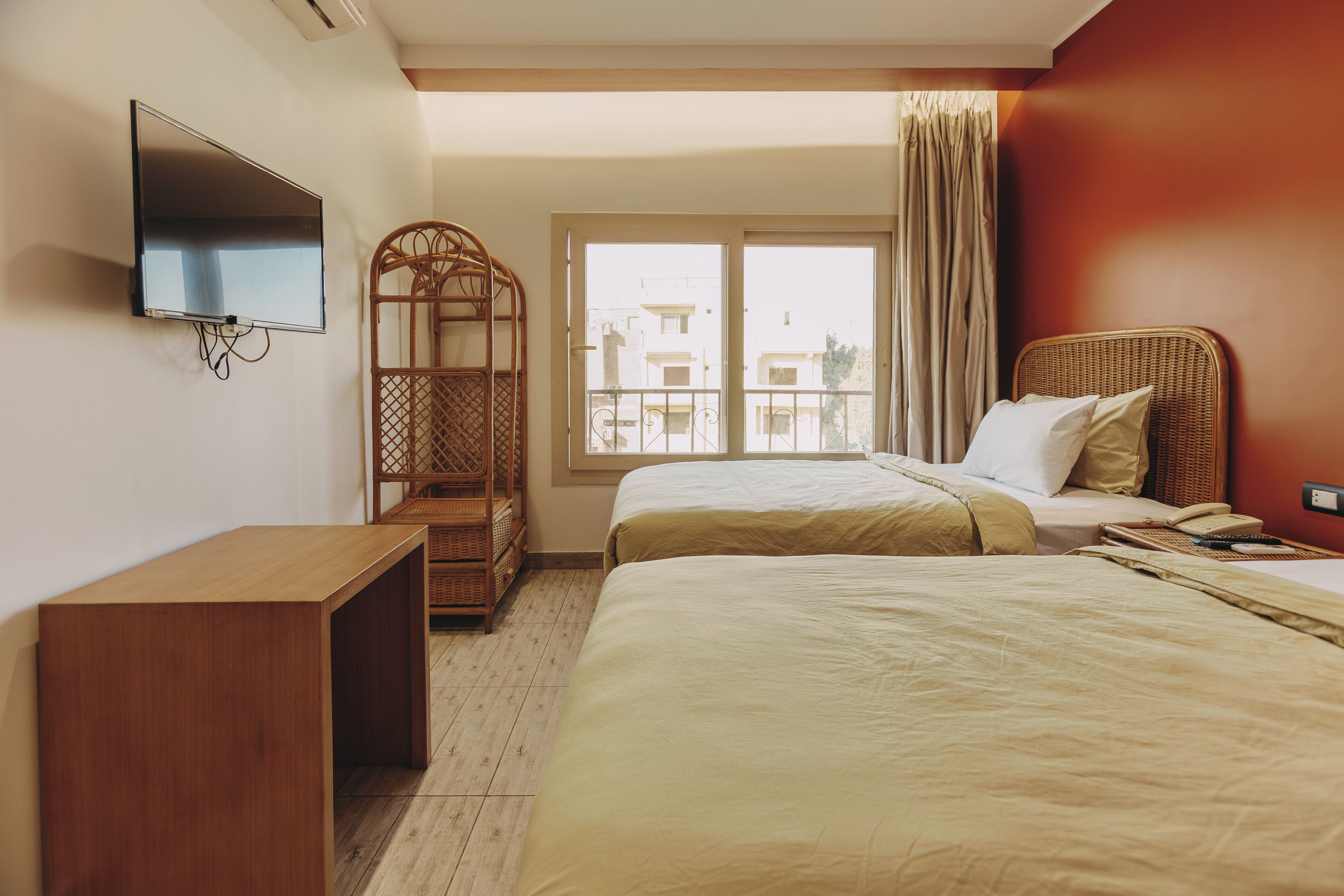 A hotel room with two beds, a TV, a desk, a window with curtains, and a balcony view.