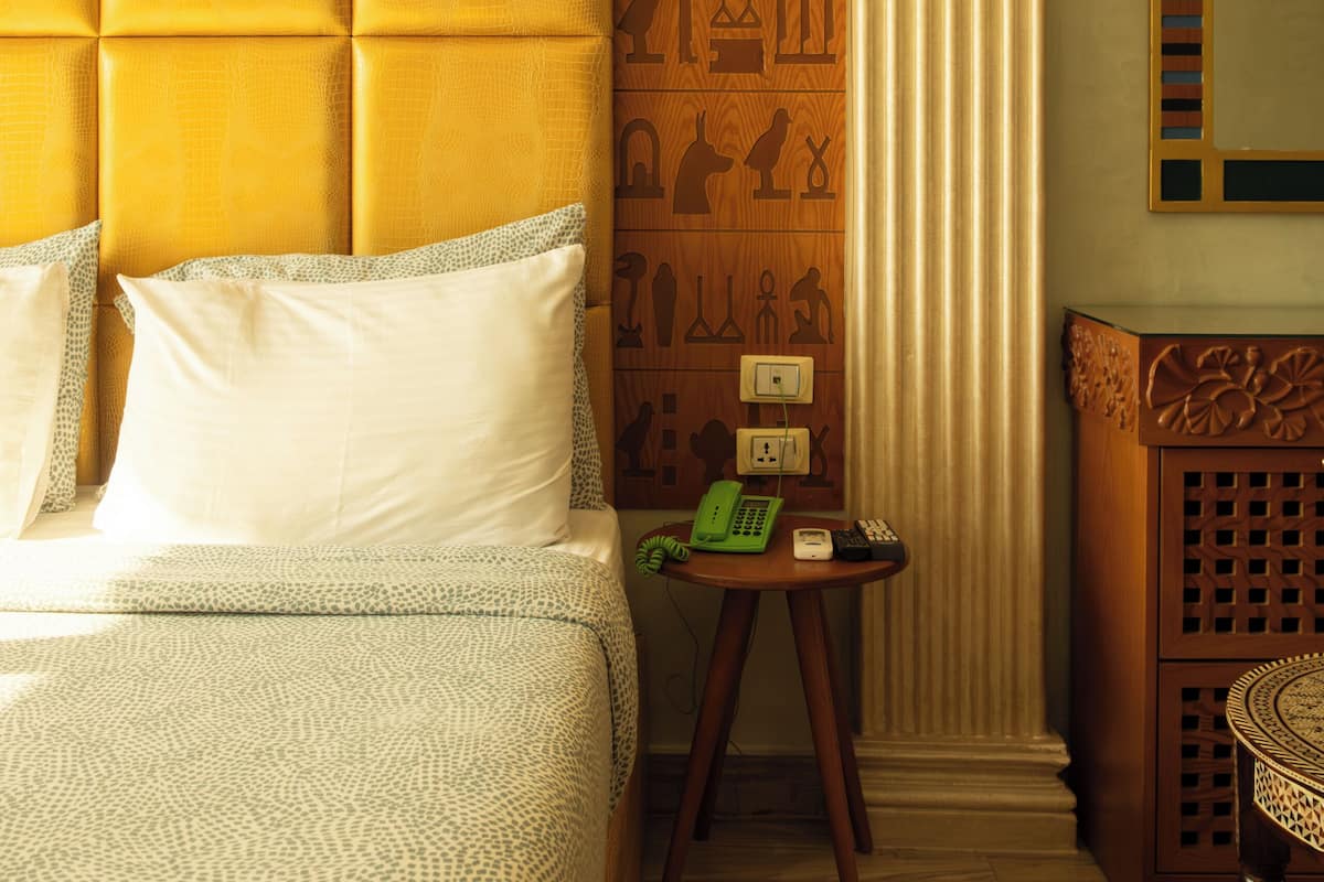 A hotel room with a bed, a small table with a telephone, a wall with Egyptian hieroglyphs, and a mirror.