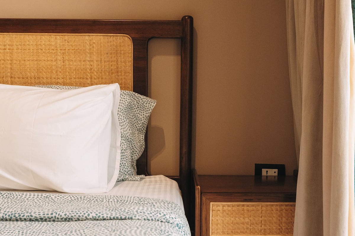 A neatly made bed with a patterned bedspread and pillows, a wooden nightstand with a power outlet, and a window with a sheer curtain.