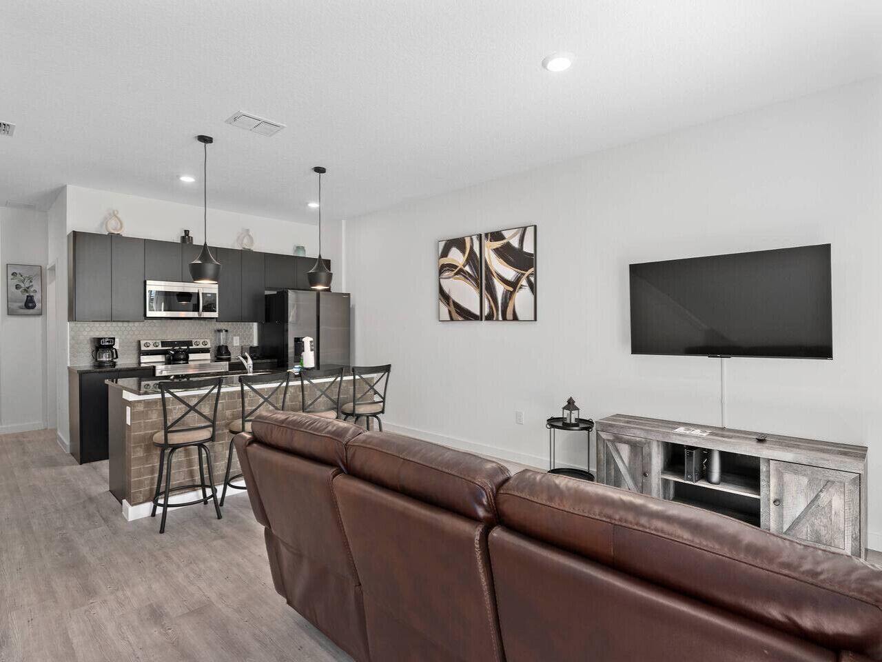 A modern living room with a brown leather sofa, a flat-screen TV mounted on the wall, and a kitchen area with a dining table and chairs.