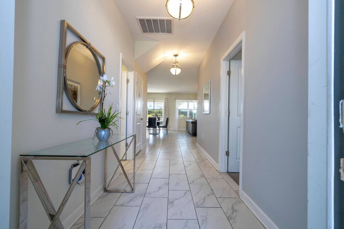 A hallway with a glass console table, a potted plant, a mirror, and a dining area in the background.