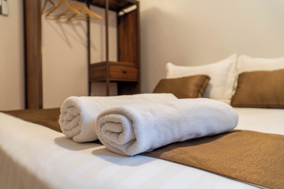 A neatly made bed with a rolled white towel, a brown throw blanket, and two brown pillows.