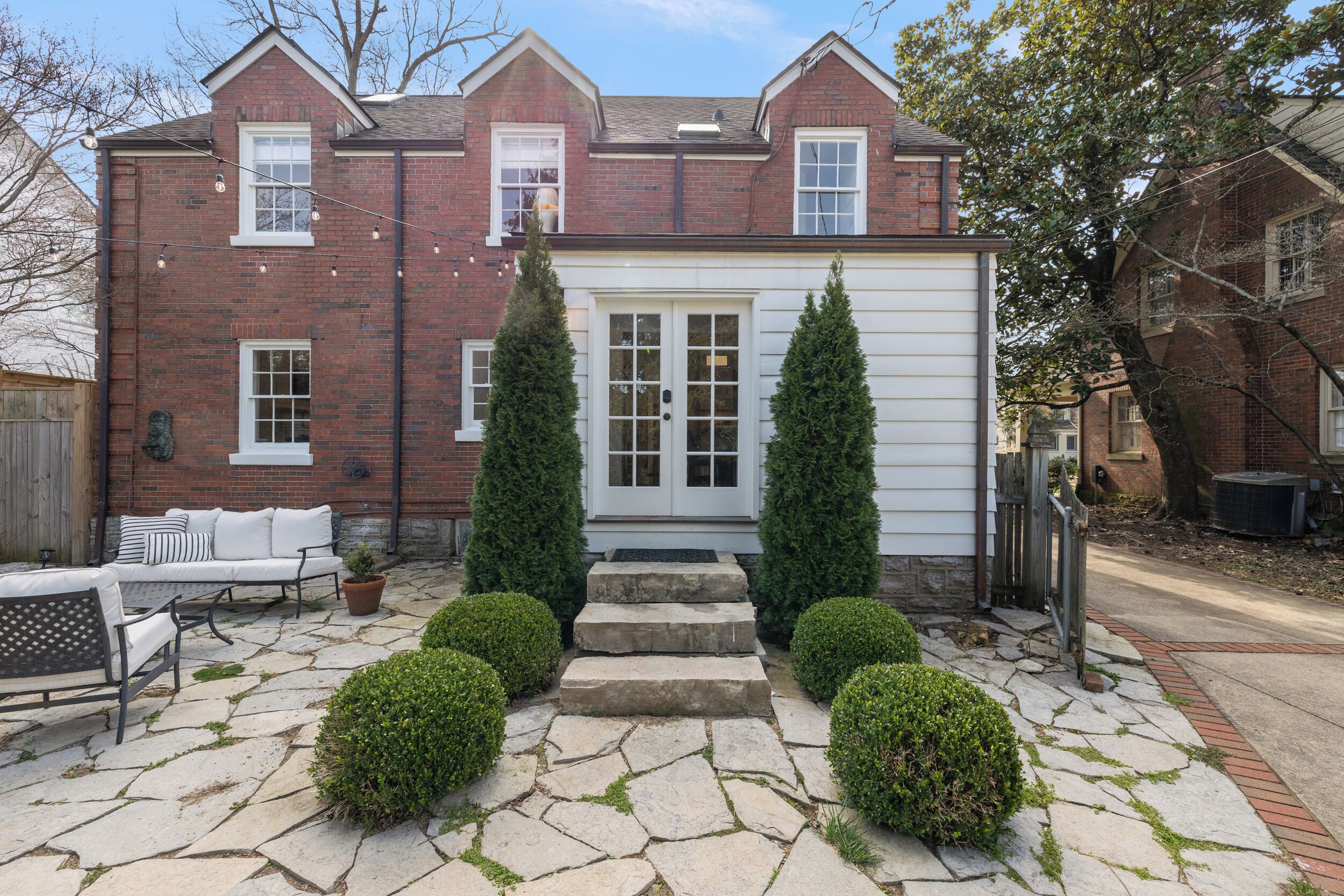 Une maison à deux étages, construite en briques, avec une porte d’entrée blanche, un petit porche avec des marches et une terrasse en pierre agrémentée de mobilier d’extérieur.