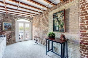 Exposed brick walls and a wooden ceiling frame this cozy space, featuring a large window, a framed painting, and a small table with a potted plant.