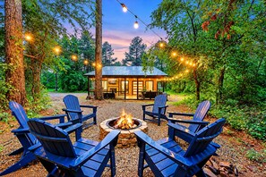 A cabin with a fire pit surrounded by Adirondack chairs, set in a forest with string lights.