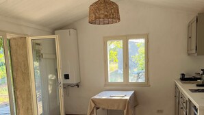A room with a tiled floor, a table with a tablecloth, a hanging light fixture, a window with a view of greenery, and a door leading outside. - Charmant Cabanon Provençal (Montfort-sur-Argens)