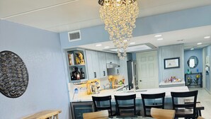 A modern dining area with a round glass table surrounded by chairs, a chandelier, and a kitchen in the background. - Panoramic Ocean Front view 3 bedroom 3 bath condo
STR License 2024-0443 (South Padre Island)