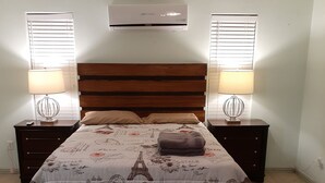 Une chambre à coucher comprenant un lit, deux tables de chevet, un ventilateur de plafond et une fenêtre avec des stores.