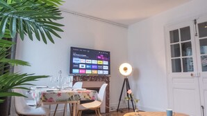 A room with a dining table set for two, a television mounted on the wall displaying various streaming services, a standing lamp, and a door with glass panels. - Paradis - Cosy Comfy 3Beds 70m² Central FastWiFi SmartTV55" PrimeVideo BBQ (Verdun)