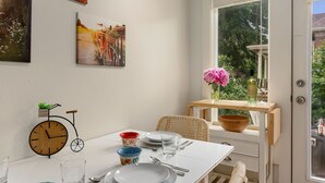 Un comedor con una mesa blanca, sillas de mimbre, un reloj de madera y una ventana con vistas a la vegetación.