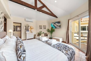 Ein Schlafzimmer mit einem großen Bett, einem Schreibtisch, einem Sessel, einer Topfpflanze, einem Gemälde und einer Glastür zum Balkon.