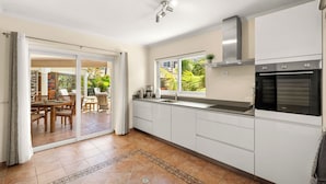 Une cuisine moderne dotée d’un four encastré, d’une fenêtre donnant sur un espace verdoyant et d’une porte-fenêtre coulissante qui mène à un coin repas extérieur.