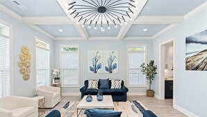 A spacious living room with a modern ceiling design, a dark blue sofa set, a white coffee table, and a large abstract painting on the wall. - The Quiet Storm - Walk to Beach - 4BR/3BA - 3 Living Areas (North Myrtle Beach)