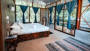 A spacious bedroom with a large bed, a ceiling fan, and multiple windows with curtains. - Kinkajoungalows (Drake Bay)