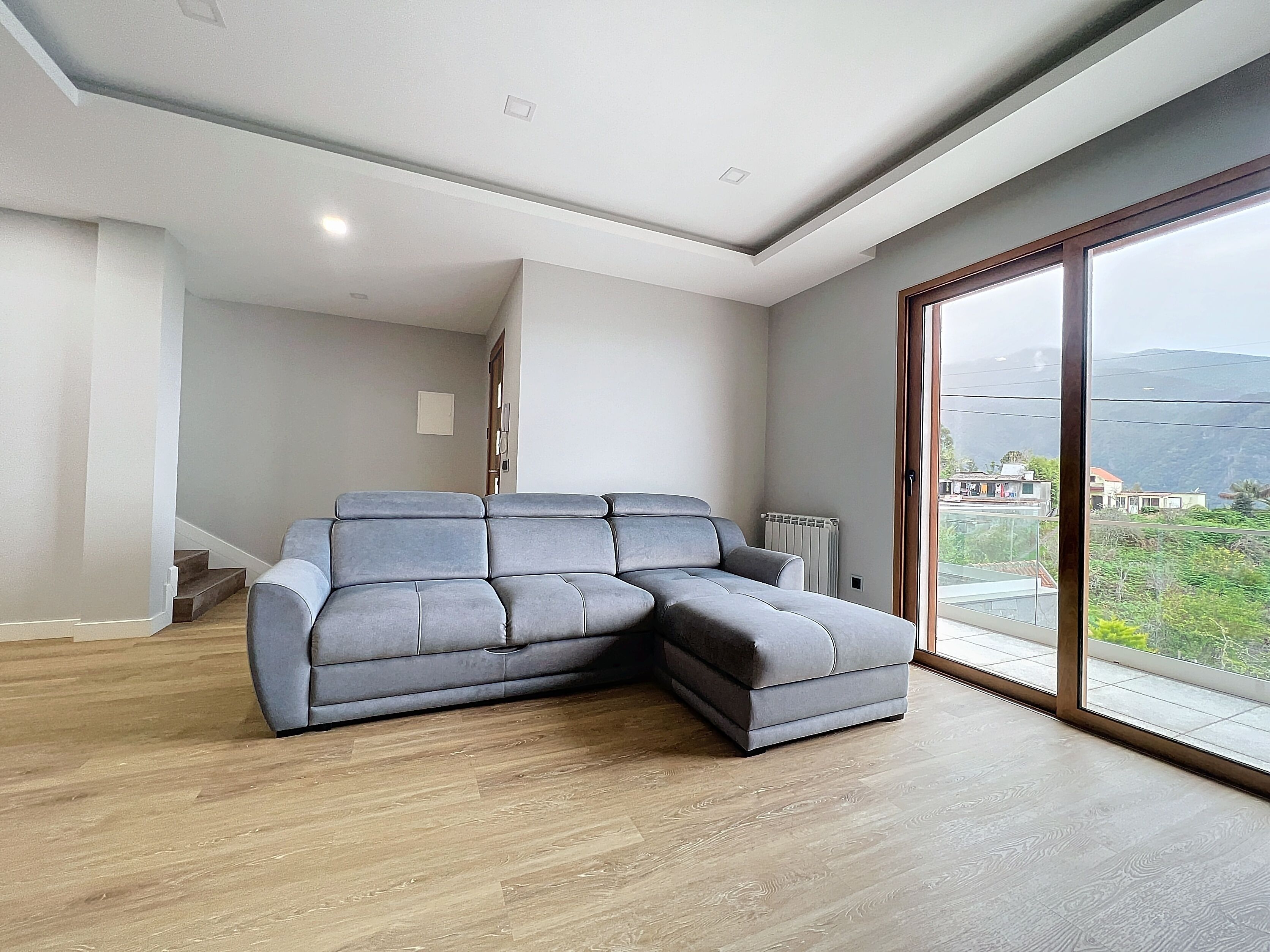 A modern living room with a sectional sofa, wooden flooring, a large sliding glass door, and a view of the outdoors.