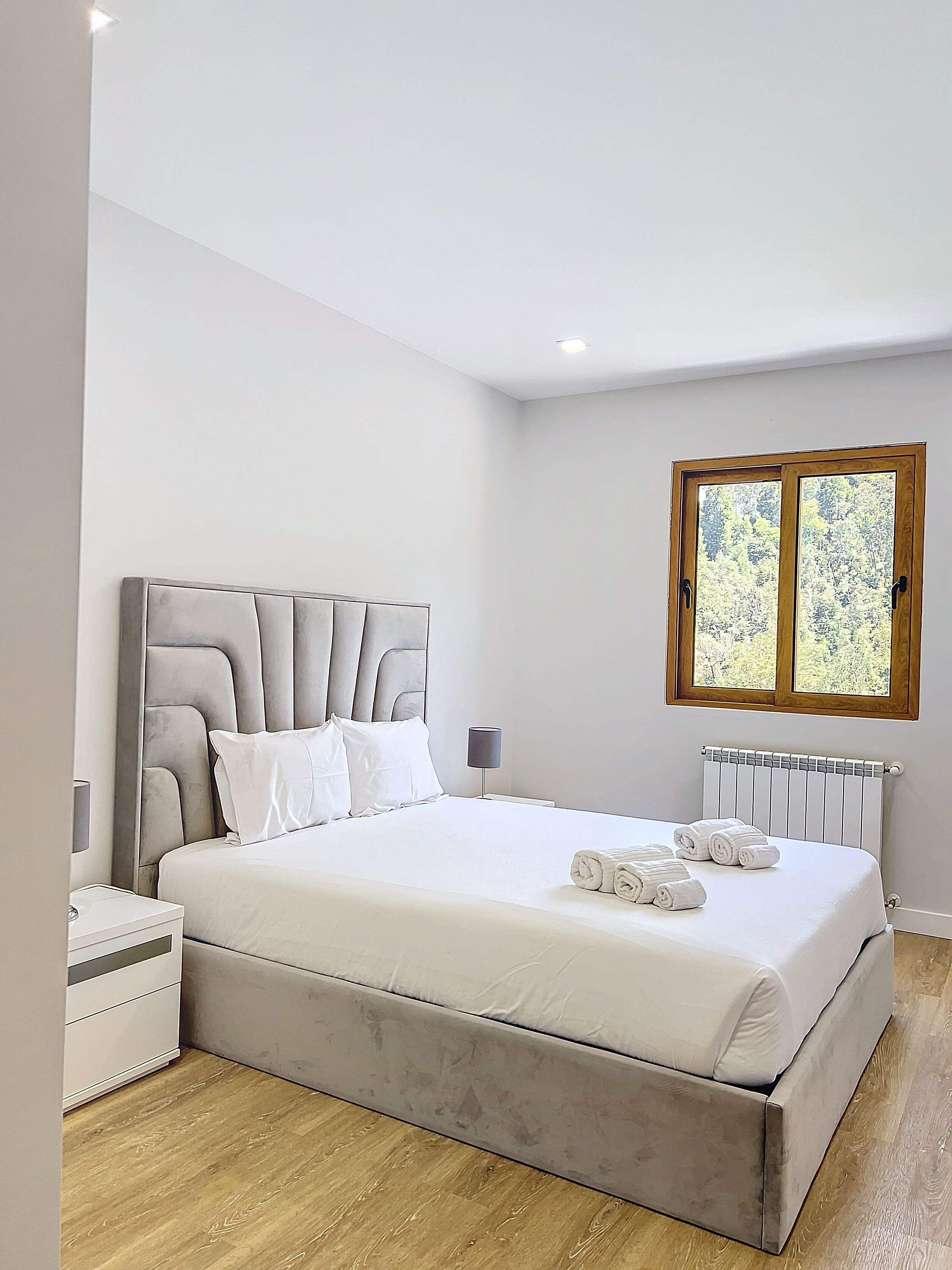 A modern bedroom with a large bed, a bedside table, a nightstand, and a window with a view of trees.