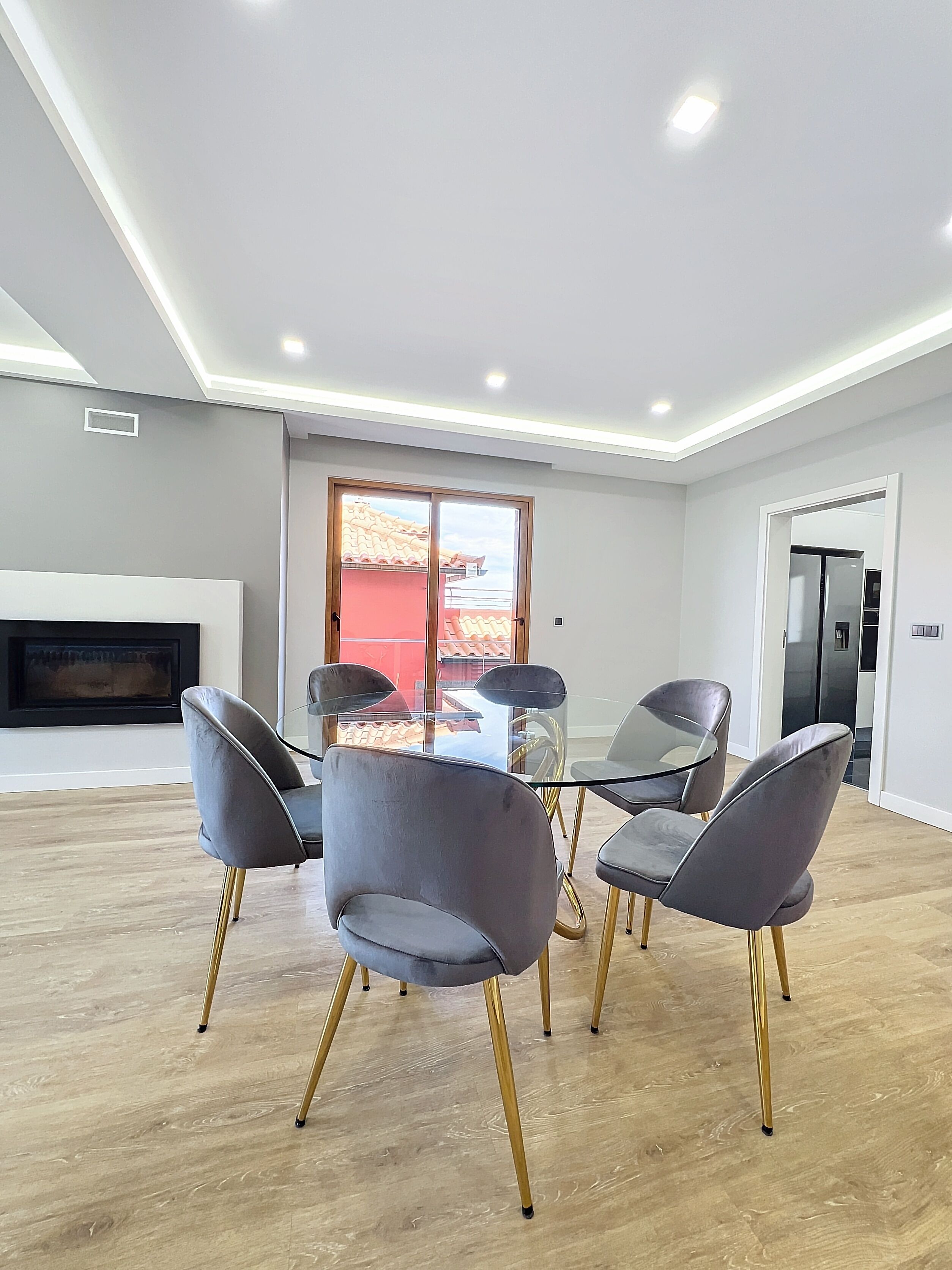 A modern dining area with a glass table and grey upholstered chairs with gold legs.