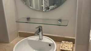 A bathroom with a round mirror, a glass shelf, a white sink, and a shower area. - Joli Studio à 2 Minutes du Centre de Monaco (Beausoleil)