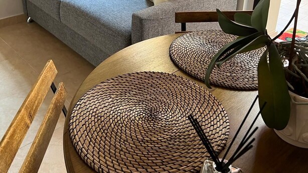 A living room with a grey sofa, a round wooden table with woven placemats, a small vase with a purple orchid, and a window with blinds. - Joli Studio à 2 Minutes du Centre de Monaco (Beausoleil)