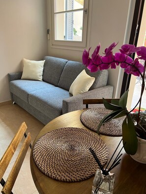 Un salon avec un canapé gris, une table ronde en bois surmontée de sets de table en fibres tressées, un petit vase contenant une orchidée violette et une fenêtre avec des stores.