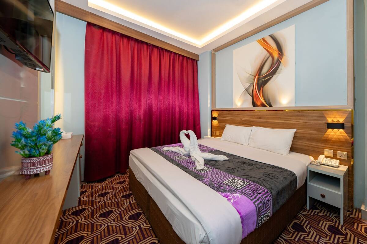 A hotel room with a bed, a nightstand, a desk, a red curtain, and a wall with a modern abstract painting.
