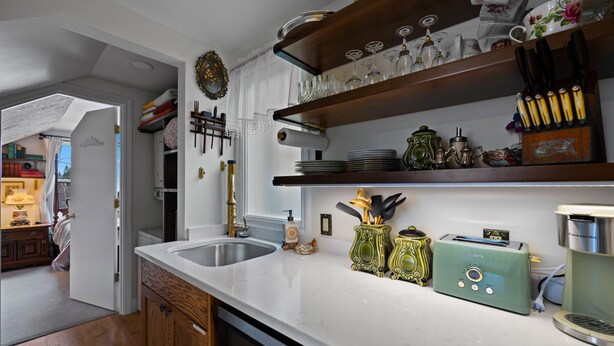 A kitchen with a white countertop, a sink, and a dishwasher. There are shelves above the countertop holding various kitchen items. - The Victorian Story at Midtown Manor (Bend)