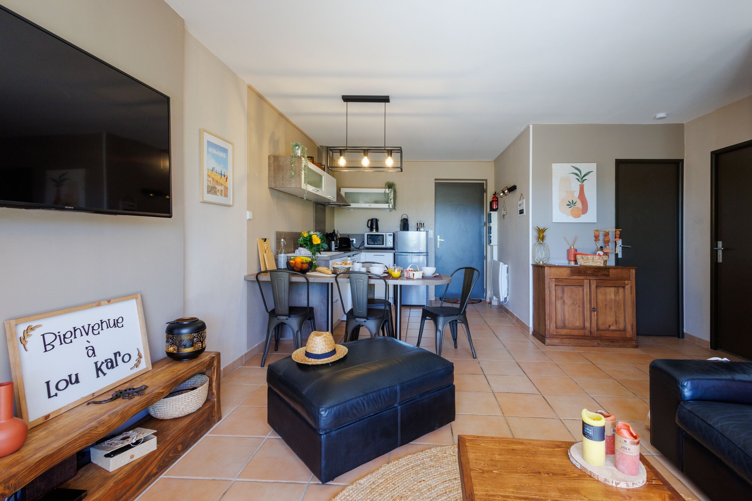 A modern living room with a black leather sofa, a wooden coffee table, and a black ottoman. There is a kitchen area with a dining table and chairs, a microwave, and a refrigerator. A TV is mounted on the wall, and there are various decorative items on shelves and tables.