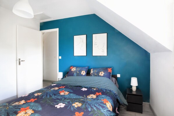 Une chambre avec un mur d’accent bleu, un lit avec une literie à motifs floraux, deux œuvres d’art murales et une table de chevet avec une lampe.