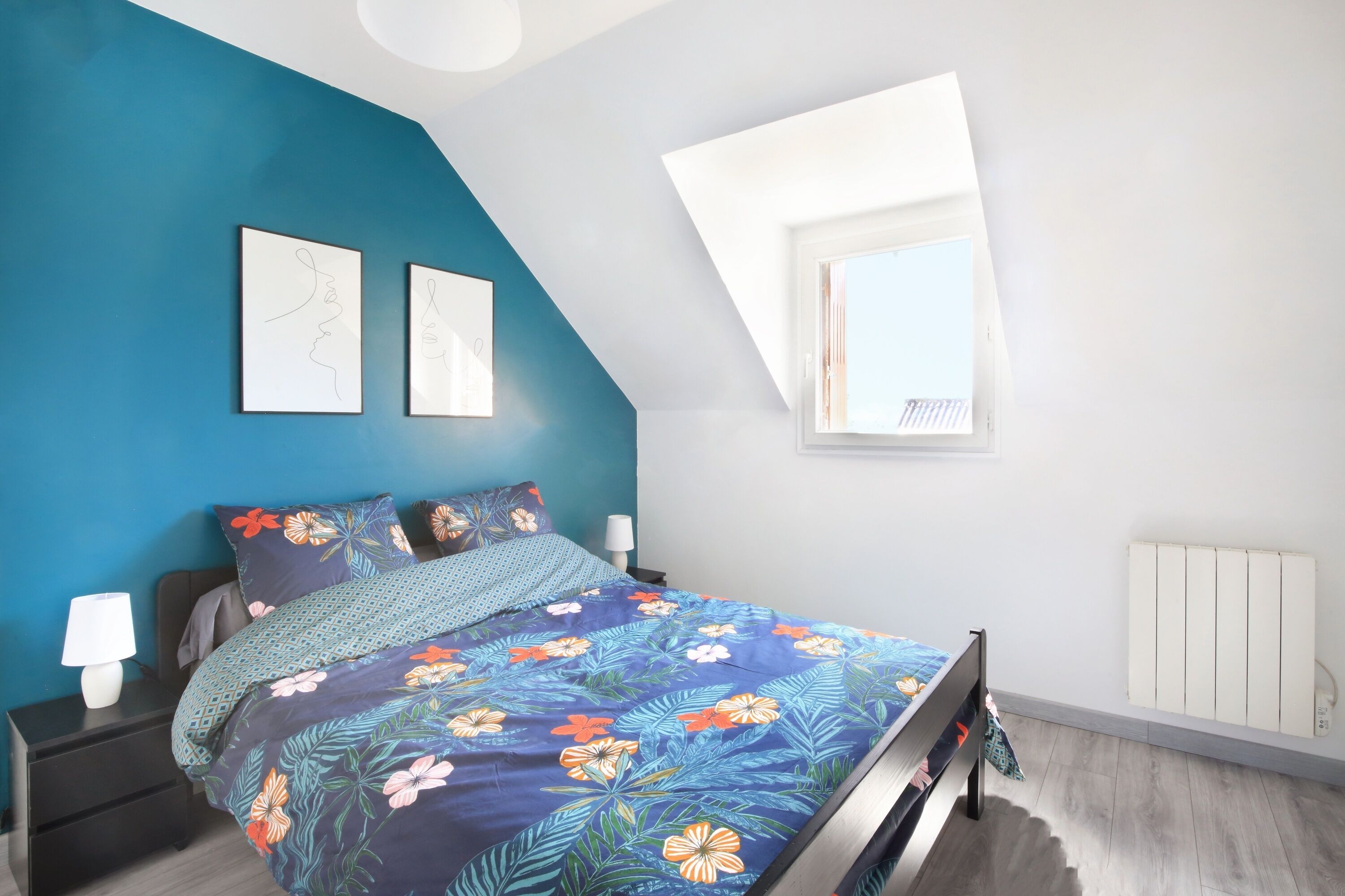 A bedroom with a sloped ceiling, a bed with a floral bedspread, two wall-mounted artworks, a bedside table with a lamp, and a window with a view of the sky.