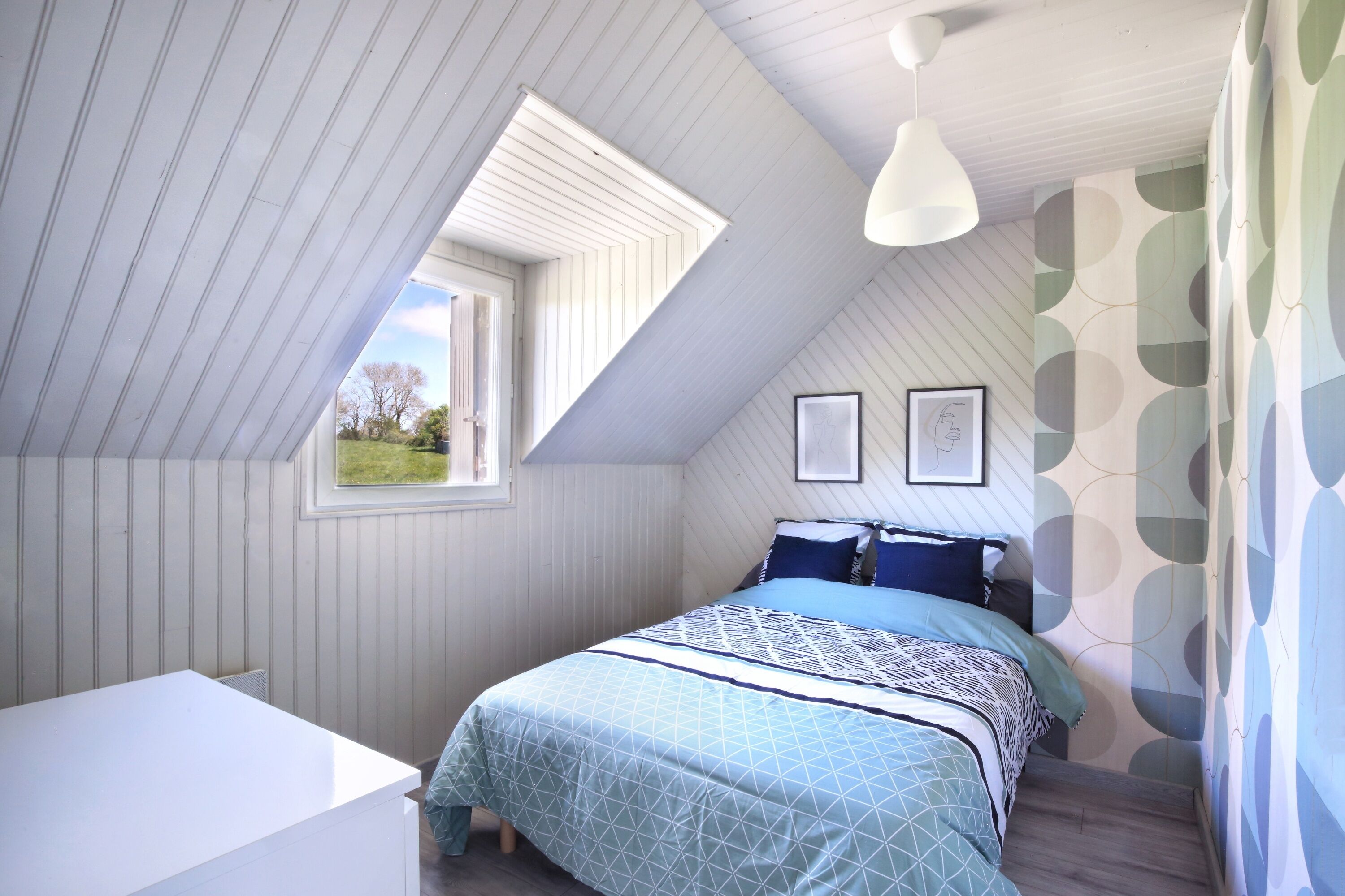 A bedroom with a sloped ceiling, a bed with patterned bedding, a window, and a wall with a geometric wallpaper design.