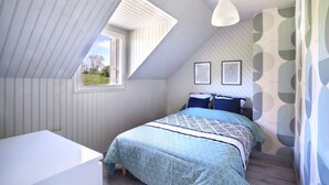 Une chambre avec un plafond en pente, un lit avec une literie à motifs, une fenêtre et un mur recouvert d’un papier peint à motifs géométriques.