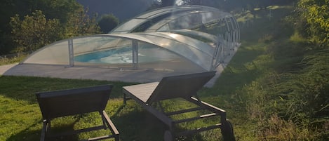 A glass-enclosed swimming pool with two wooden chairs in a grassy area, surrounded by hills and trees.