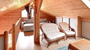 Ein geräumiger Loft mit Holzbalken, einer Couch, zwei Korbstühlen und Blick ins Freie.
