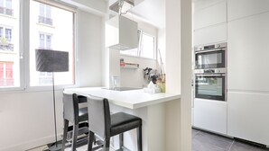 Een moderne keuken met een wit aanrecht, twee zwarte barkrukken, een inbouwoven en een raam dat natuurlijk licht biedt.
