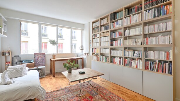 Een kamer met een bed, een tafel met een plant, een boekenkast en een groot raam.